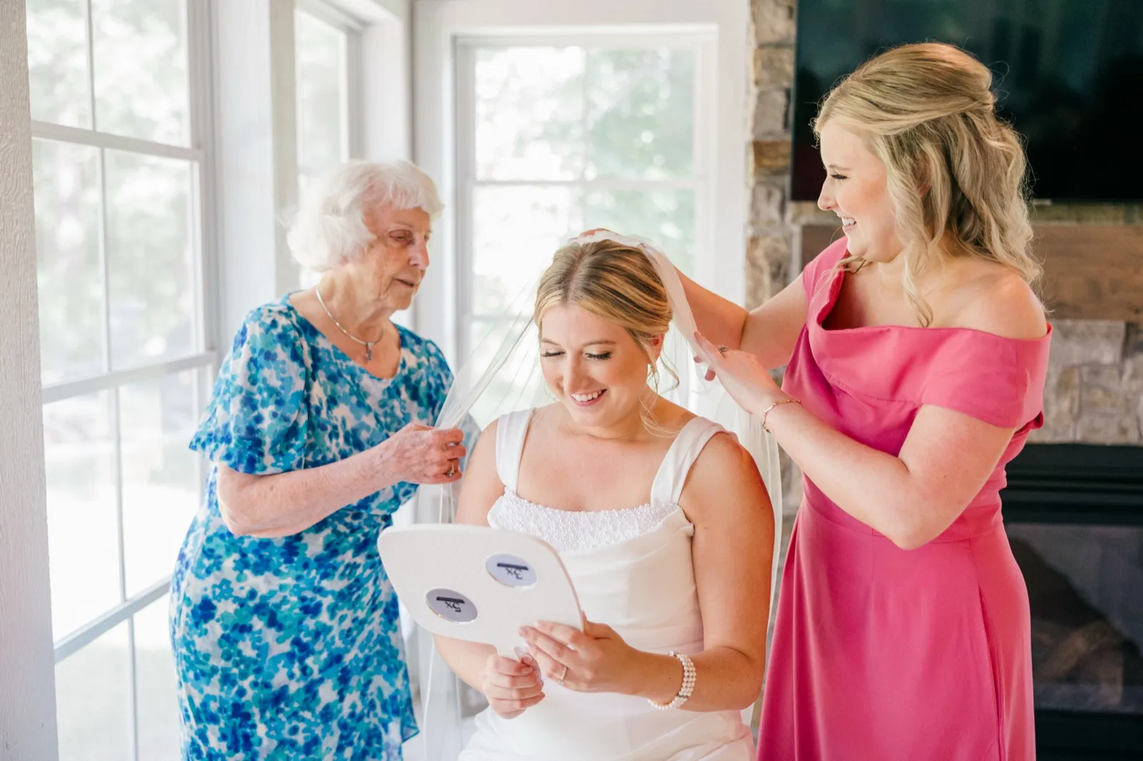 The bride looks down at a hand mirror smiling while her grandmother and a bridesmaid help adjust her veil — Tim Larsen Photography, Brainerd Lakes MN