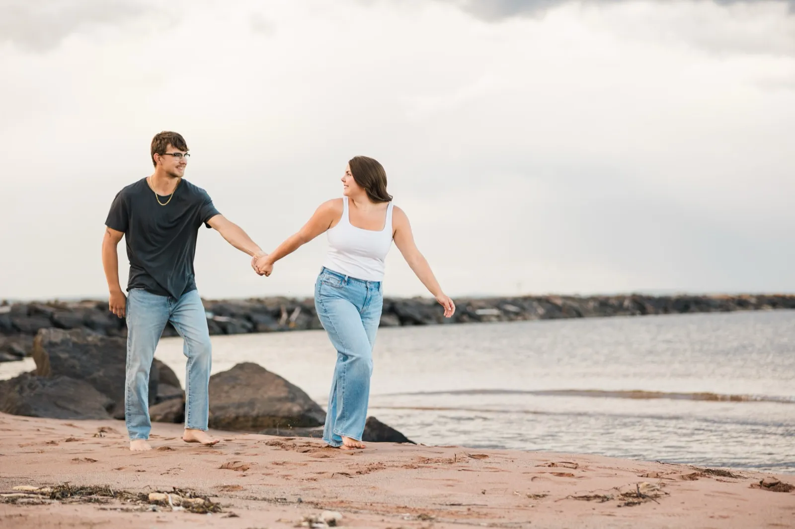 Jacklyn leads Reeden by the hand along the sandy beach, glancing back at him, the breakwater behind them — Tim Larsen Photography