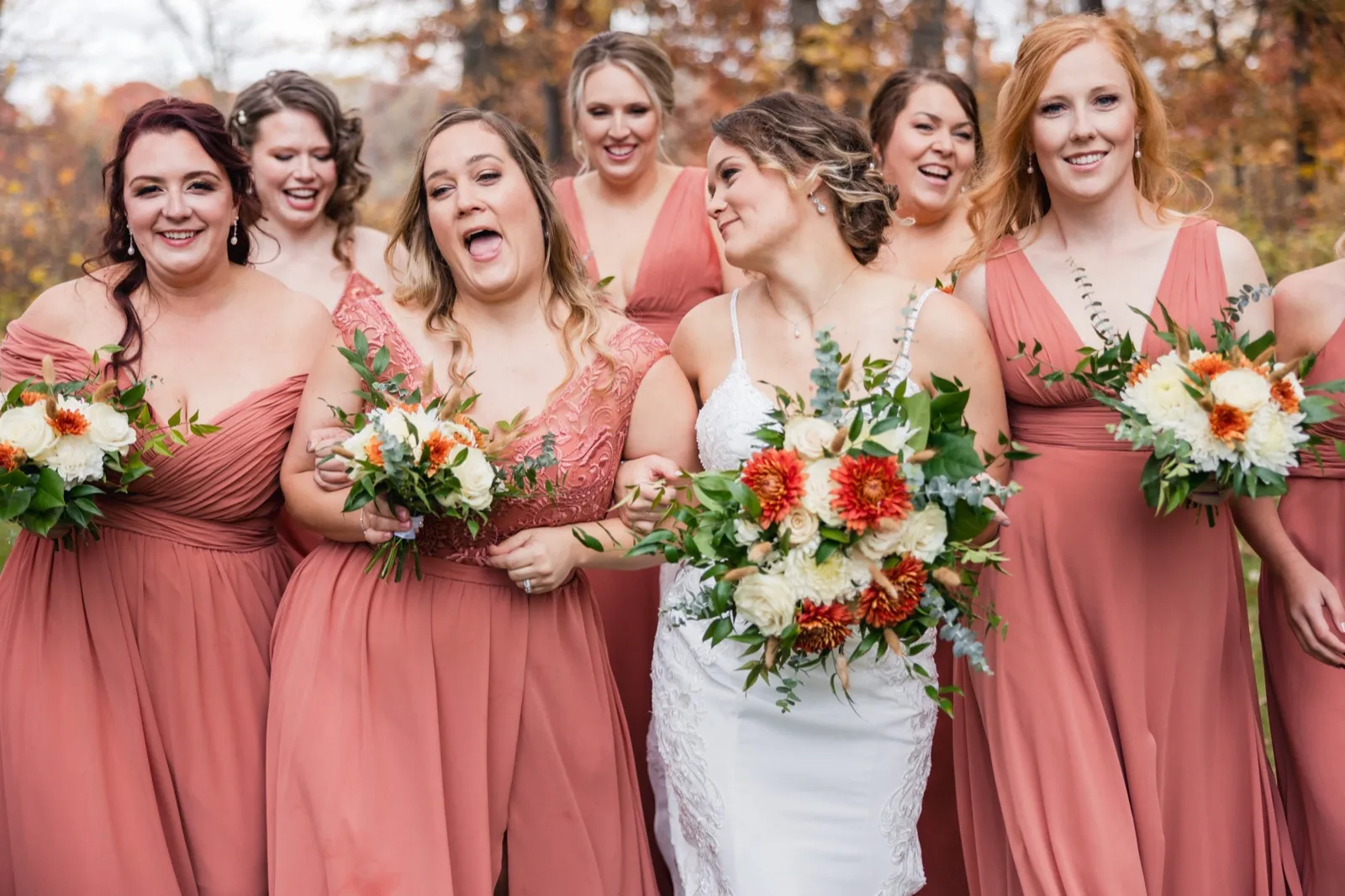 The bride and her bridesmaids in coral dresses laugh together while holding autumn-toned bouquets in front of fall foliage — Tim Larsen Photography, Brainerd Lakes MN