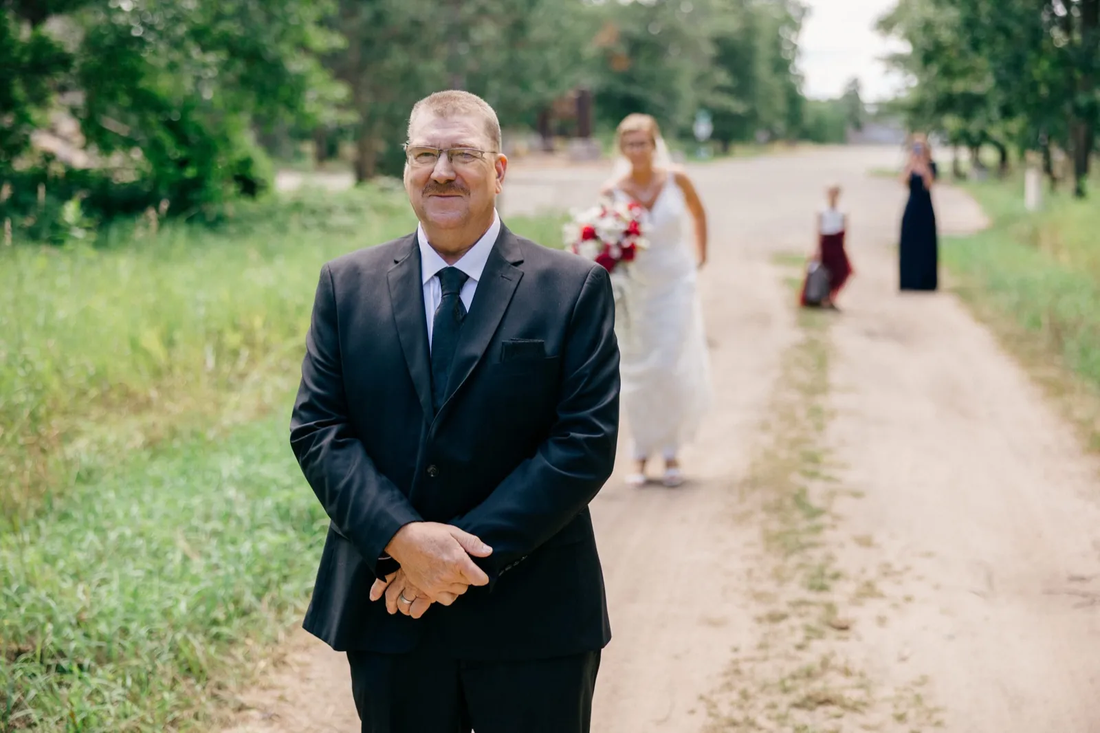 Kim's father in a dark suit waits on a dirt road as the bride approaches from behind, holding her bouquet — Tim Larsen Photography, Brainerd Lakes MN