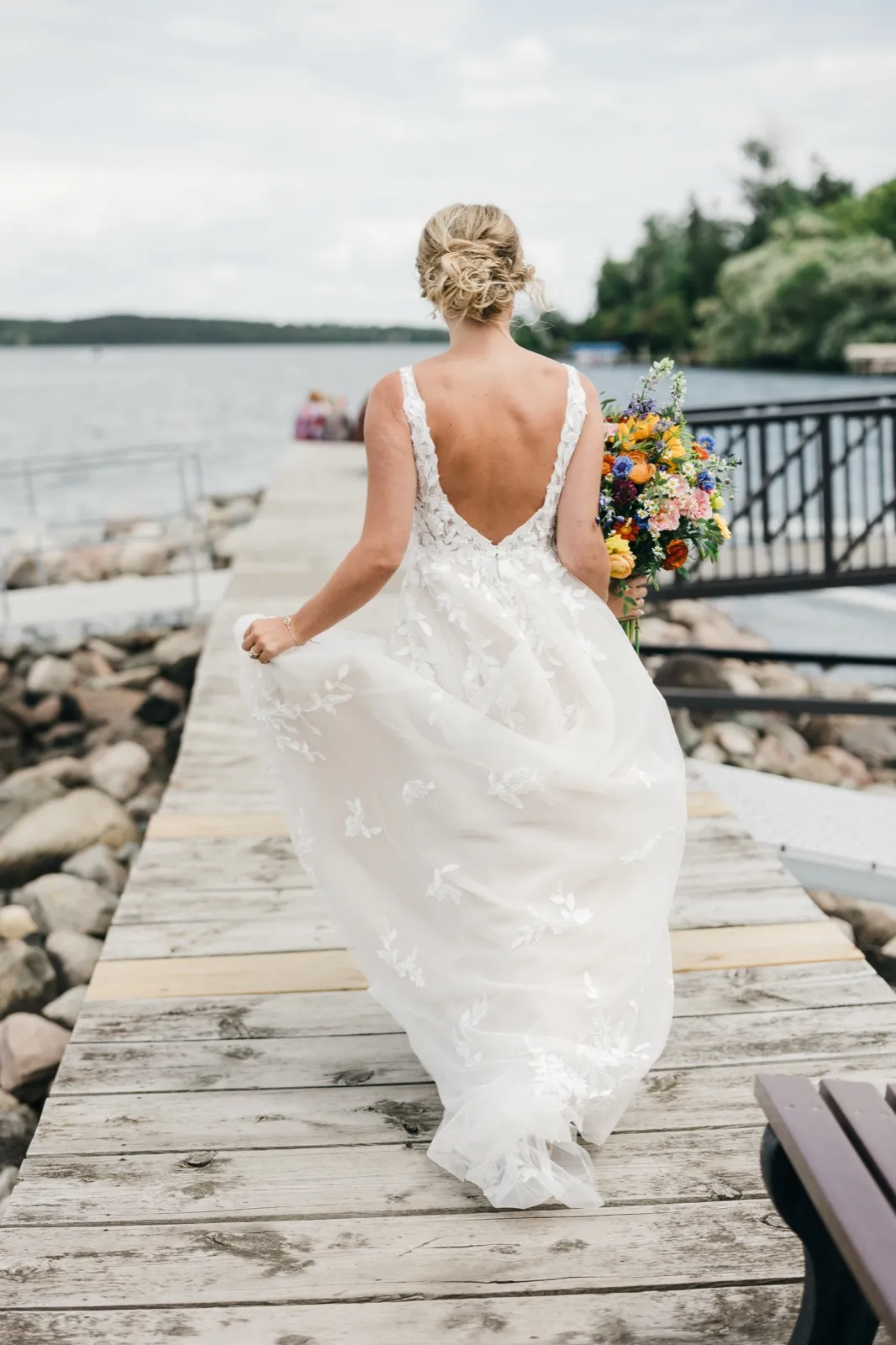 The bride walks away from the camera down a wooden dock toward the lake, holding a colorful bouquet at her side and her lace train trailing behind her. — Tim Larsen Photography, Brainerd Lakes MN