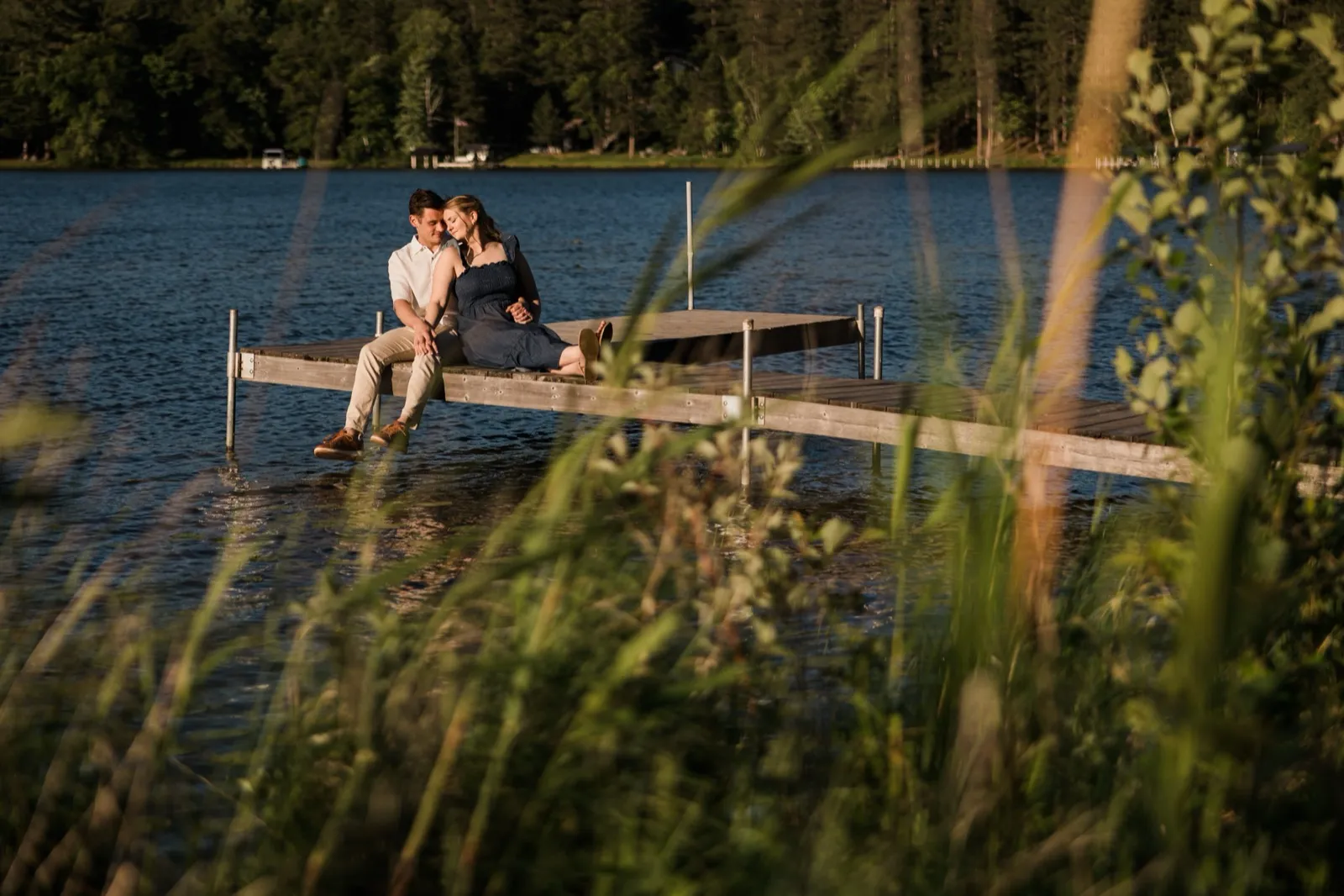 An engaged couple sit together at the end of a dock with their legs dangling over Roy Lake at Grand View Lodge — Tim Larsen Photography