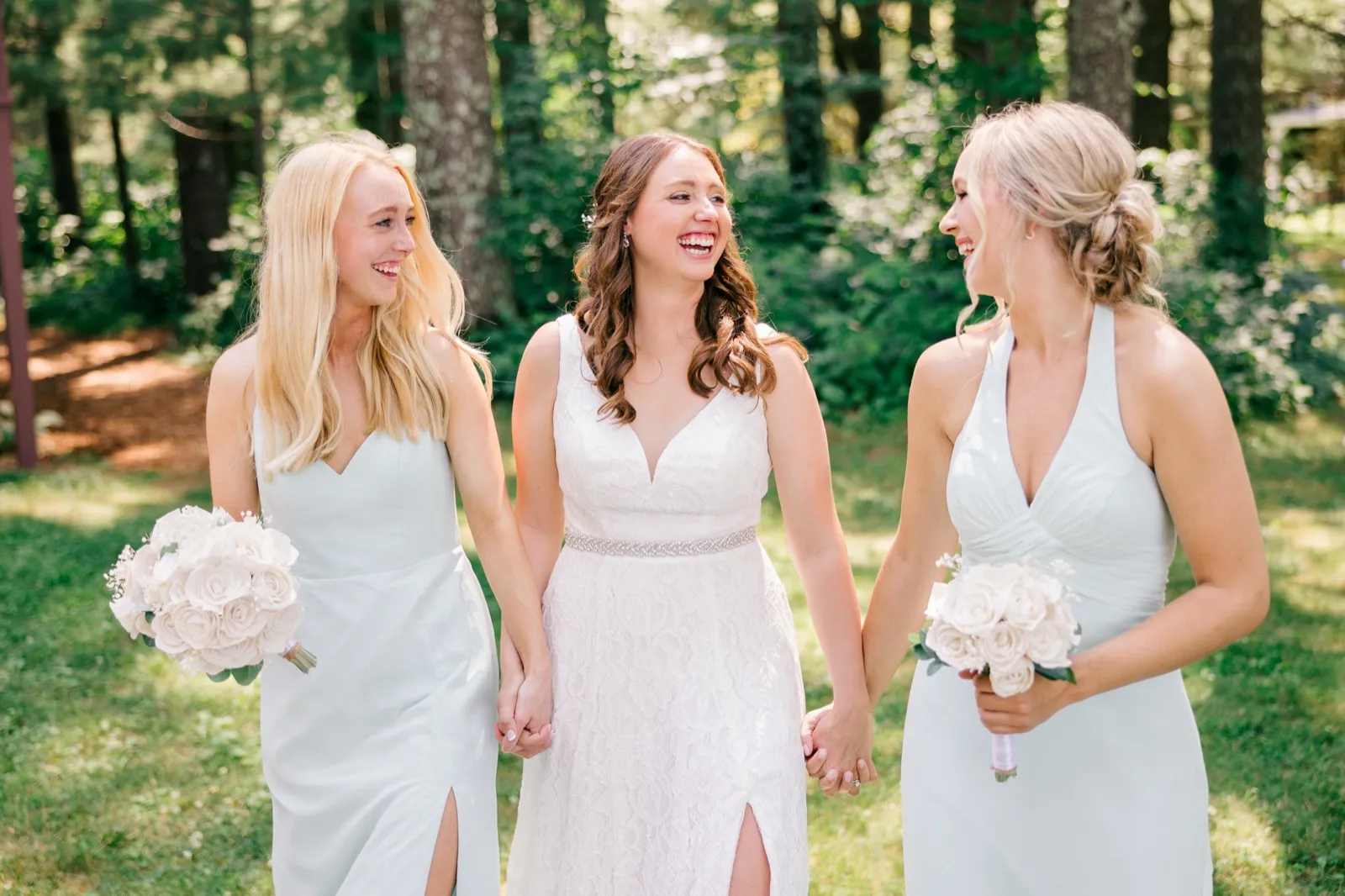 The bride laughs with two bridesmaids in pale blue dresses as they hold hands and white bouquets on a sunlit wooded lawn. — Tim Larsen Photography, Brainerd Lakes MN
