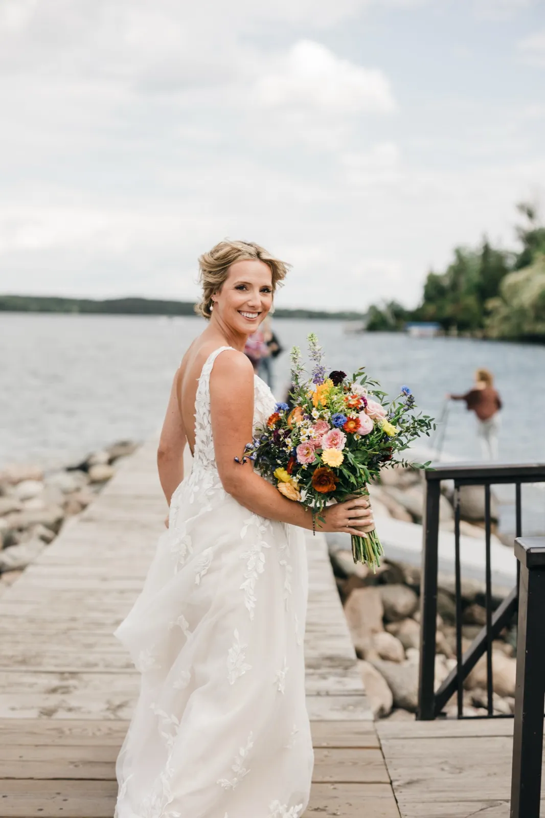 The bride glances over her shoulder with a wide smile while holding a large, colorful wildflower bouquet on a wooden dock with the lake behind her. — Tim Larsen Photography, Brainerd Lakes MN