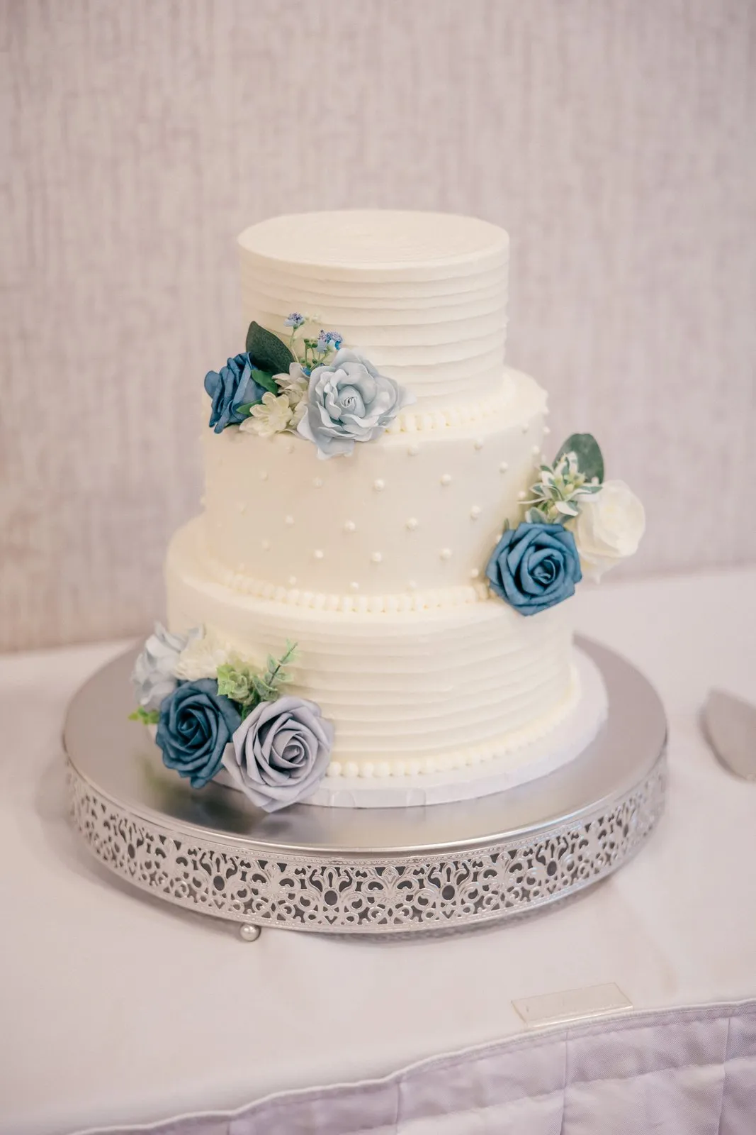 A three-tier white wedding cake with horizontal pearl-bead detailing on a wooden stand, dusty-blue roses and eucalyptus accents along the base — the reception cake at Grand View Lodge — Tim Larsen Photography, Brainerd Lakes MN