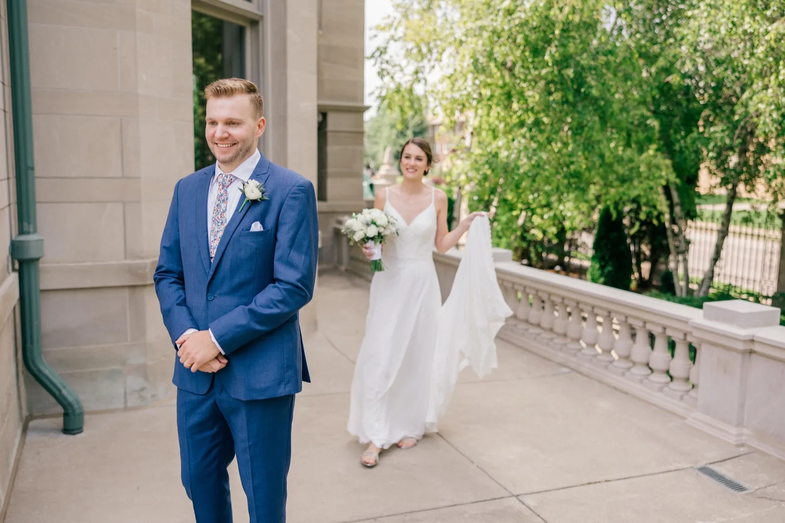 The groom stands waiting on a stone terrace as the bride approaches behind him, holding her bouquet and lifting the train of her dress. — Tim Larsen Photography, Minneapolis MN
