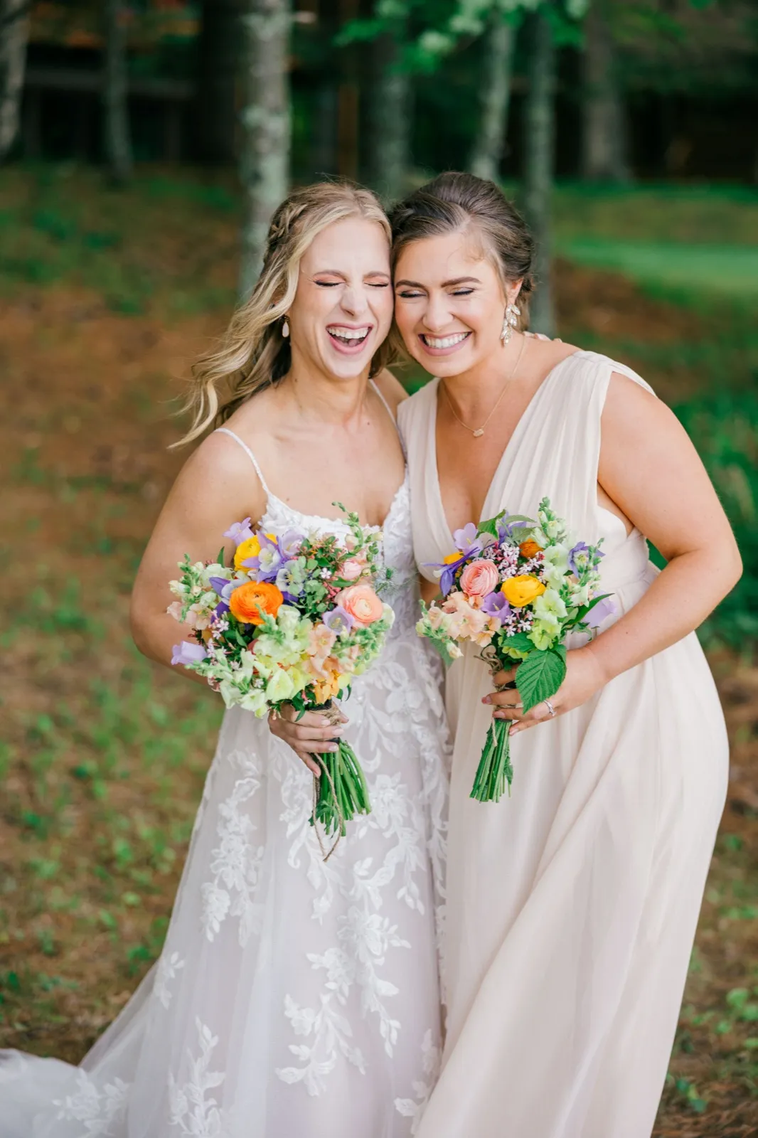 The bride and a bridesmaid lean cheek-to-cheek laughing wide-mouthed while holding colorful wildflower bouquets in a wooded clearing — Tim Larsen Photography, Brainerd Lakes MN