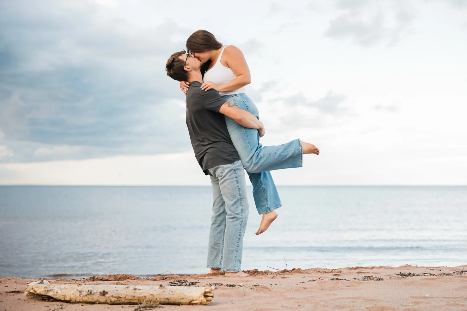 Reeden lifts Jacklyn off the sand into a kiss, Lake Superior and a wide sky behind them — Tim Larsen Photography