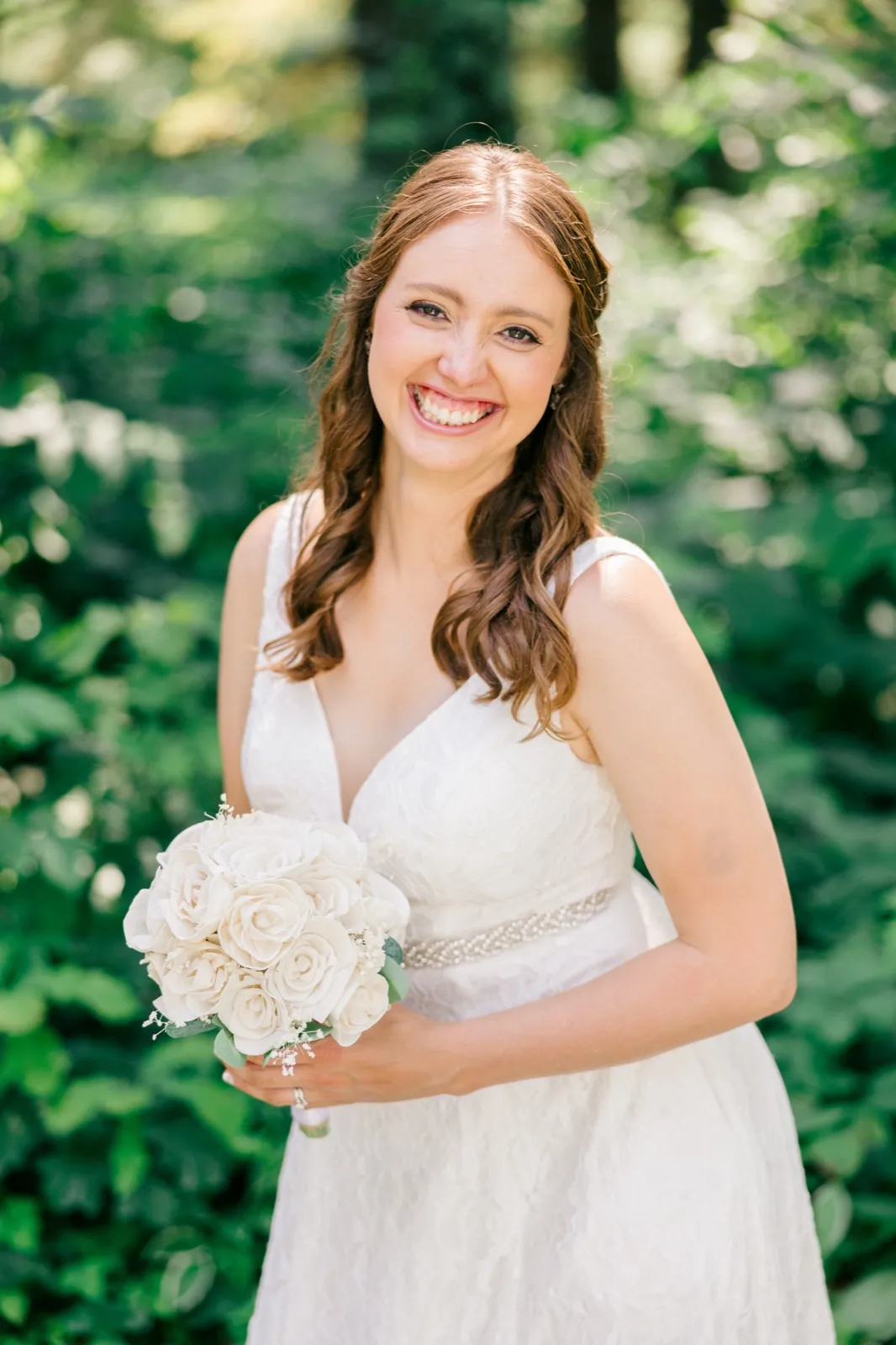 The bride smiles broadly at the camera while holding a white rose bouquet, surrounded by a soft green forest backdrop. — Tim Larsen Photography, Brainerd Lakes MN