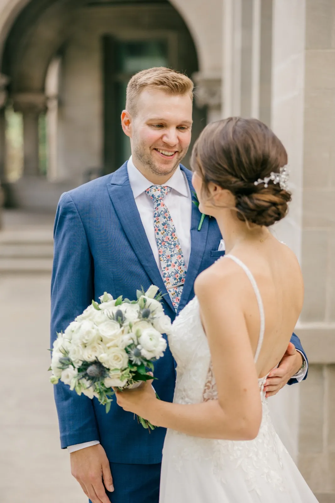 The groom smiles broadly at his bride during a first look in front of a stone archway, while she holds a white bouquet with thistle accents. — Tim Larsen Photography, Minneapolis MN