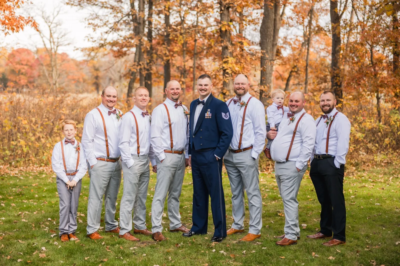The groom in military dress uniform stands centered with eight groomsmen and a young boy wearing rust-colored suspenders and bow ties in front of an autumn forest — Tim Larsen Photography, Brainerd Lakes MN