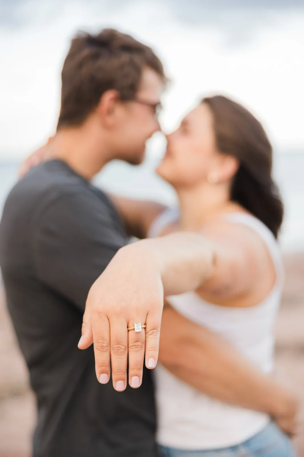Jacklyn holds her hand out to show her engagement ring while she and Reeden share a soft, blurred near-kiss behind it — Tim Larsen Photography