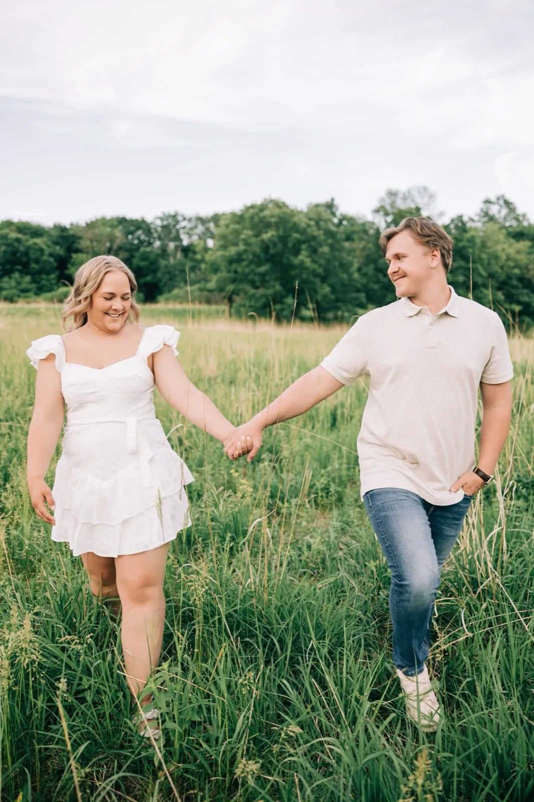 Abby and Anthony walk hand in hand through a waist-high meadow at golden hour, the tree line soft behind them — Tim Larsen Photography