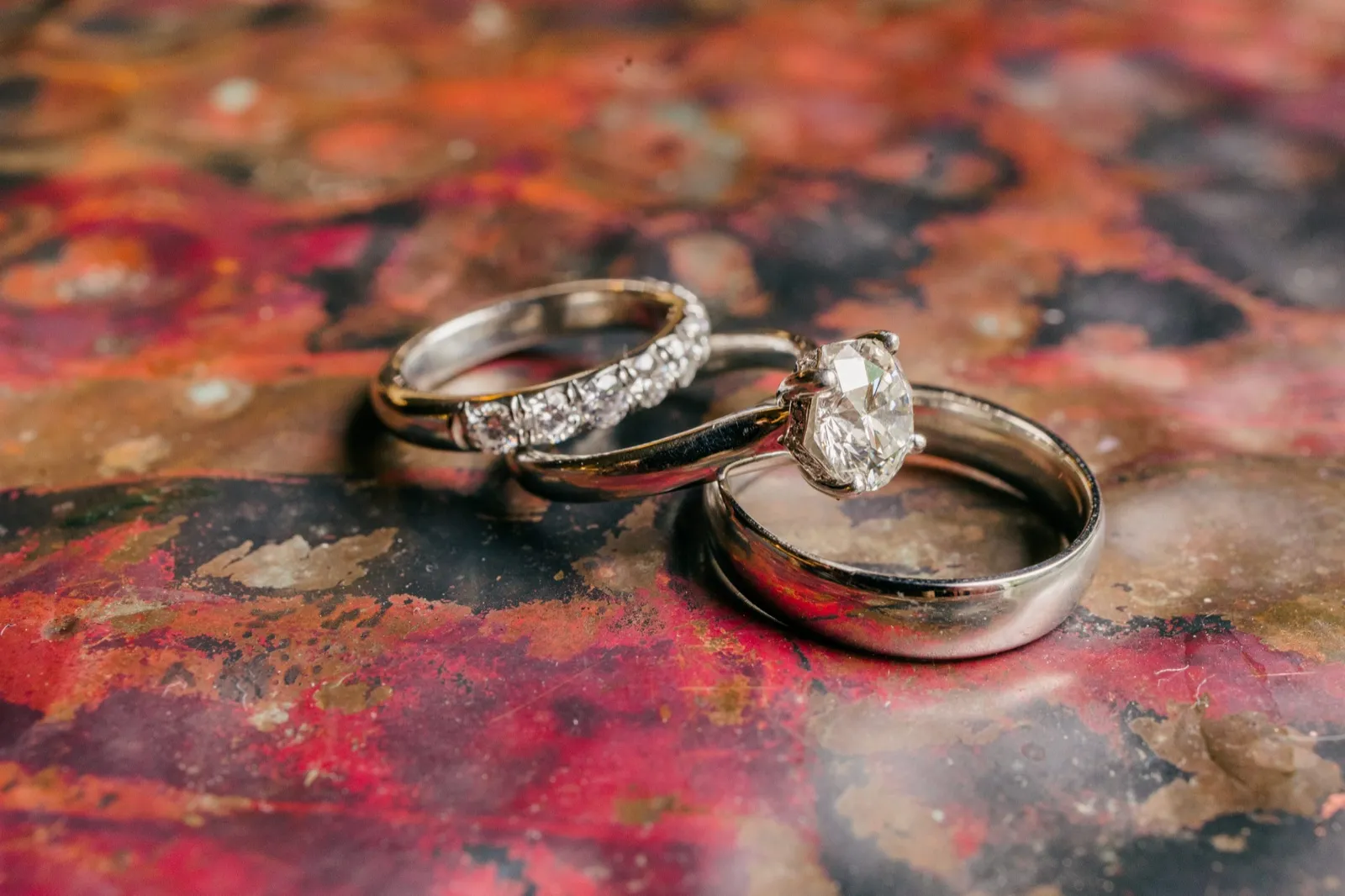 Three wedding rings including a diamond solitaire engagement ring rest on a weathered red and gold painted surface in window light — Tim Larsen Photography, Brainerd Lakes MN