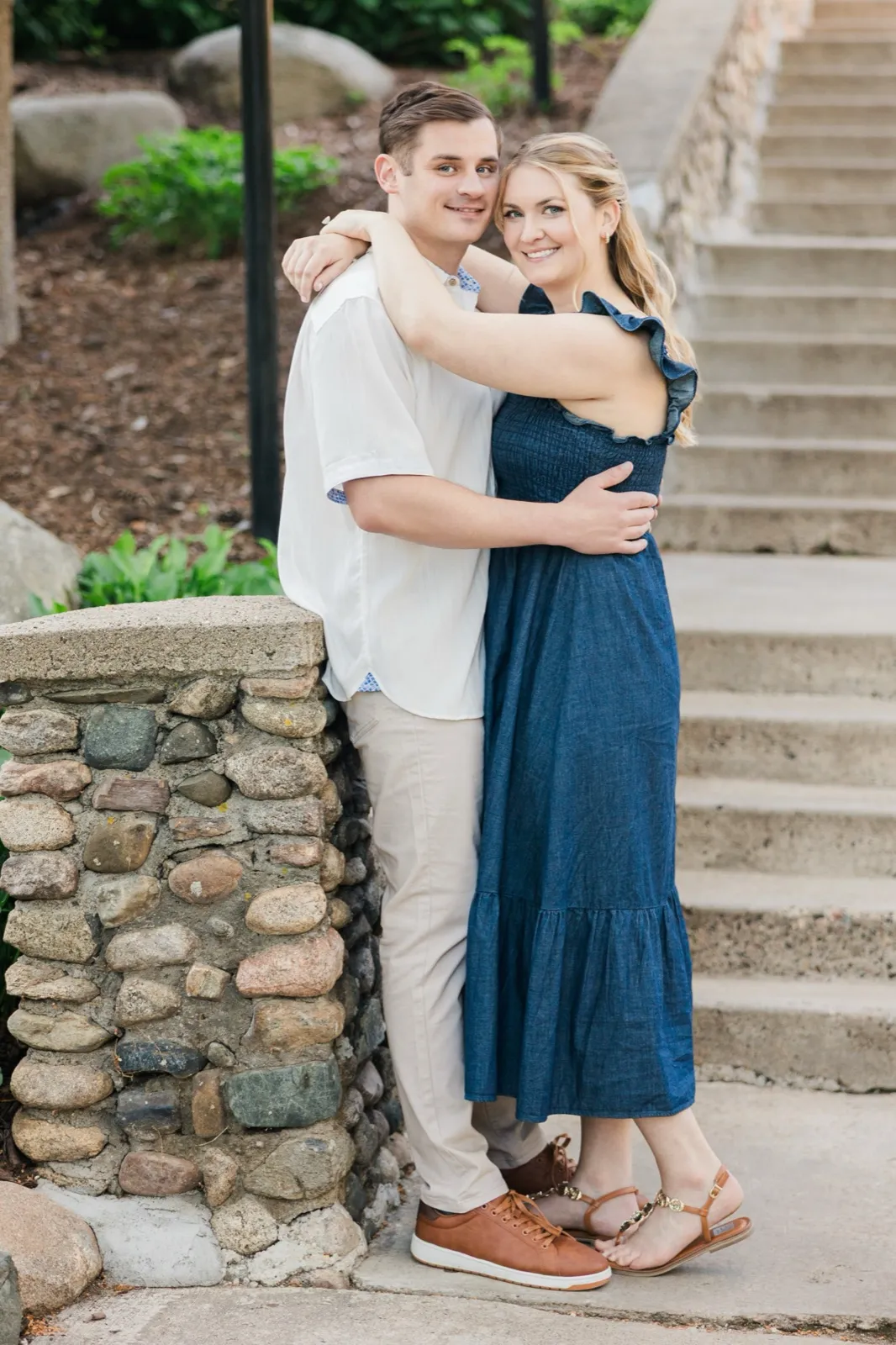 An engaged couple embrace against a river-rock stone pillar beside the staircase at Grand View Lodge, smiling at the camera — Tim Larsen Photography