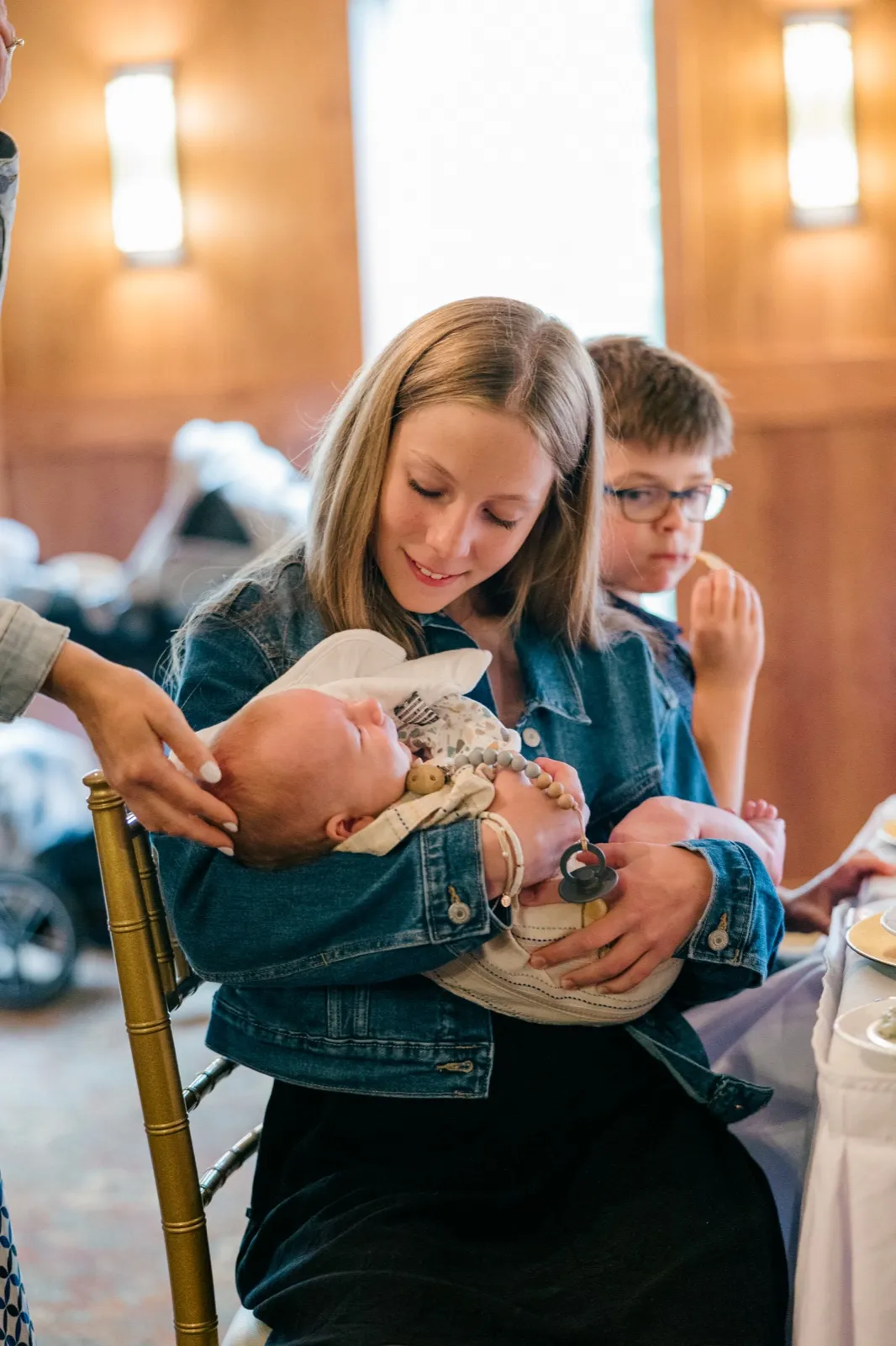 A young girl in a denim jacket smiles down at an infant cradled in her arms while a boy in glasses sits beside her at a Grand View Lodge reception table — Tim Larsen Photography, Brainerd Lakes MN