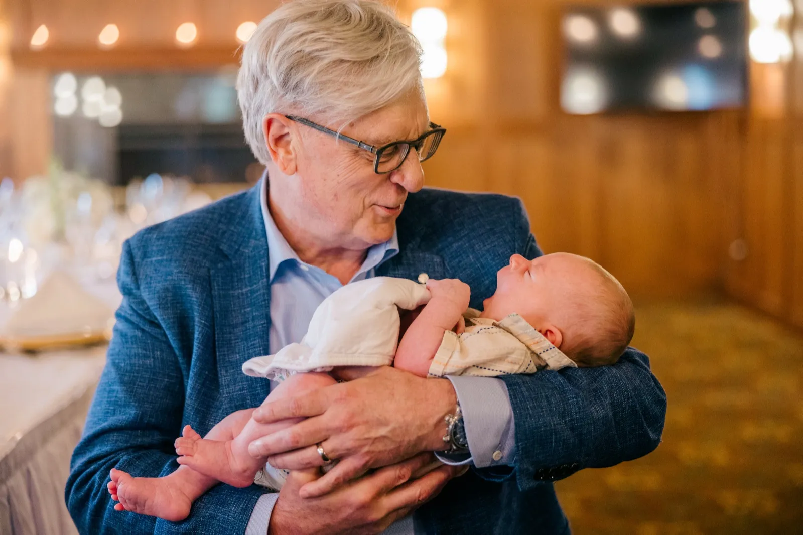 An older man in a blue blazer cradles an infant in his arms in a softly lit reception room at Grand View Lodge, the two looking at each other quietly — Tim Larsen Photography, Brainerd Lakes MN