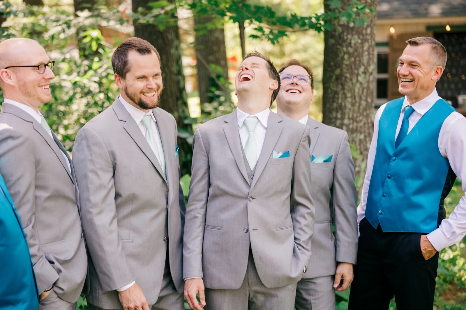 The groom laughs with his head thrown back while four groomsmen in gray suits and teal accents laugh around him in a wooded yard. — Tim Larsen Photography, Brainerd Lakes MN