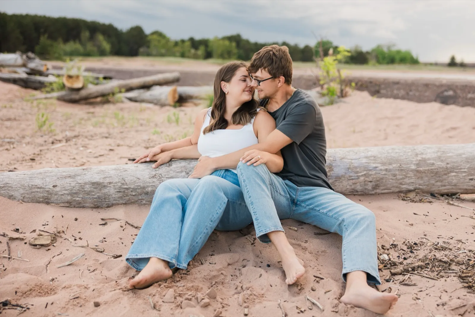 Reeden and Jacklyn lean into each other on a big driftwood log, dune grass and the tree line of Wisconsin Point behind them — Tim Larsen Photography