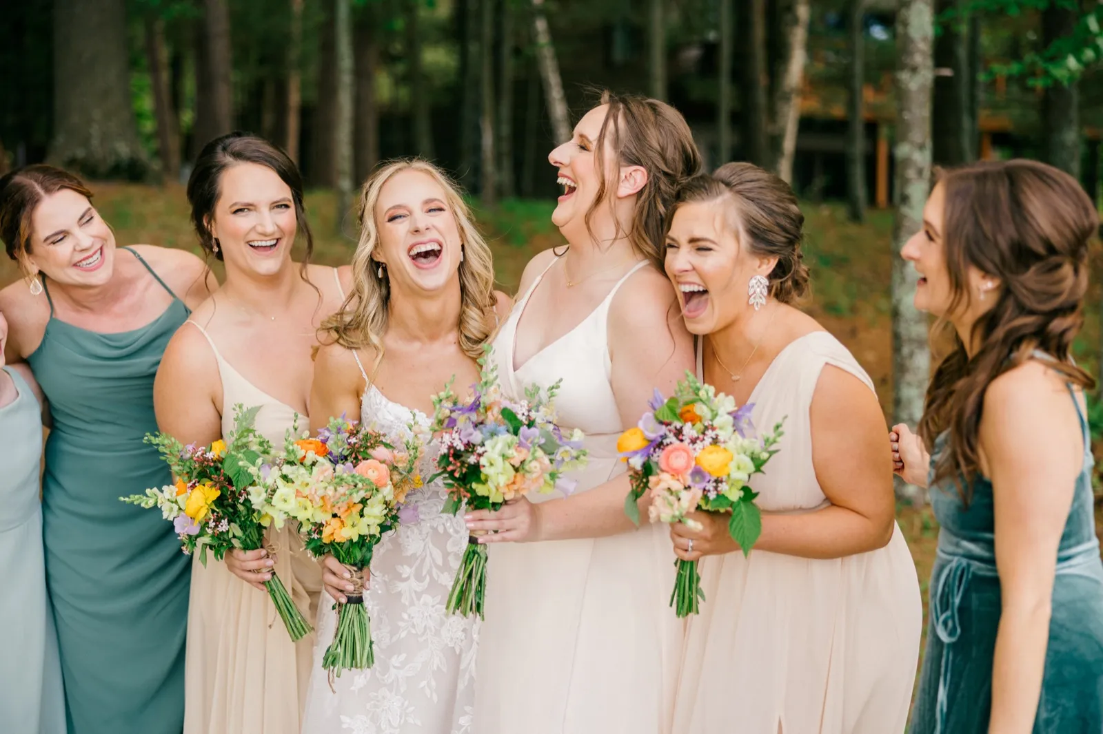 The bride and her five bridesmaids laugh together holding colorful spring bouquets on a wooded lawn outside the cabin — Tim Larsen Photography, Brainerd Lakes MN