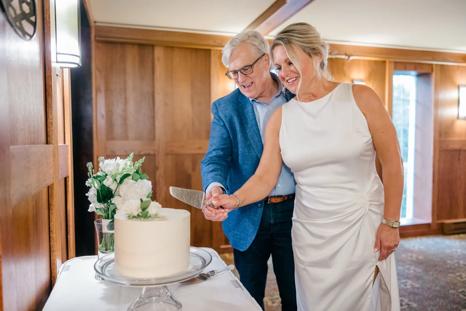 The bride and groom laugh together as they cut a small white wedding cake topped with white florals in a wood-paneled dining room — Tim Larsen Photography, Brainerd Lakes MN