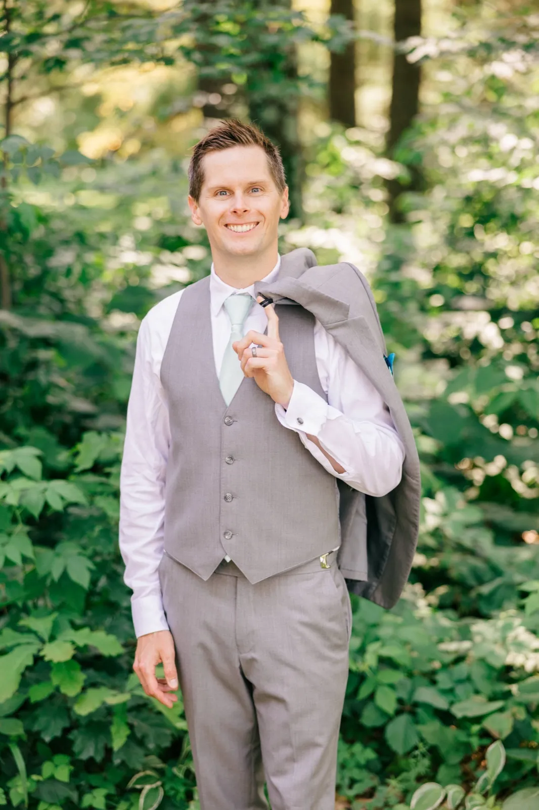 The groom smiles at the camera while holding his suit jacket over his shoulder, standing in a sunlit forest clearing. — Tim Larsen Photography, Brainerd Lakes MN