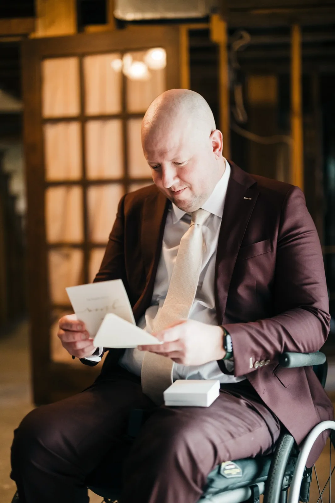 The groom, seated in his wheelchair and wearing a burgundy suit, reads a handwritten note from a small card while afternoon window light falls across him. — Tim Larsen Photography, Brainerd Lakes MN