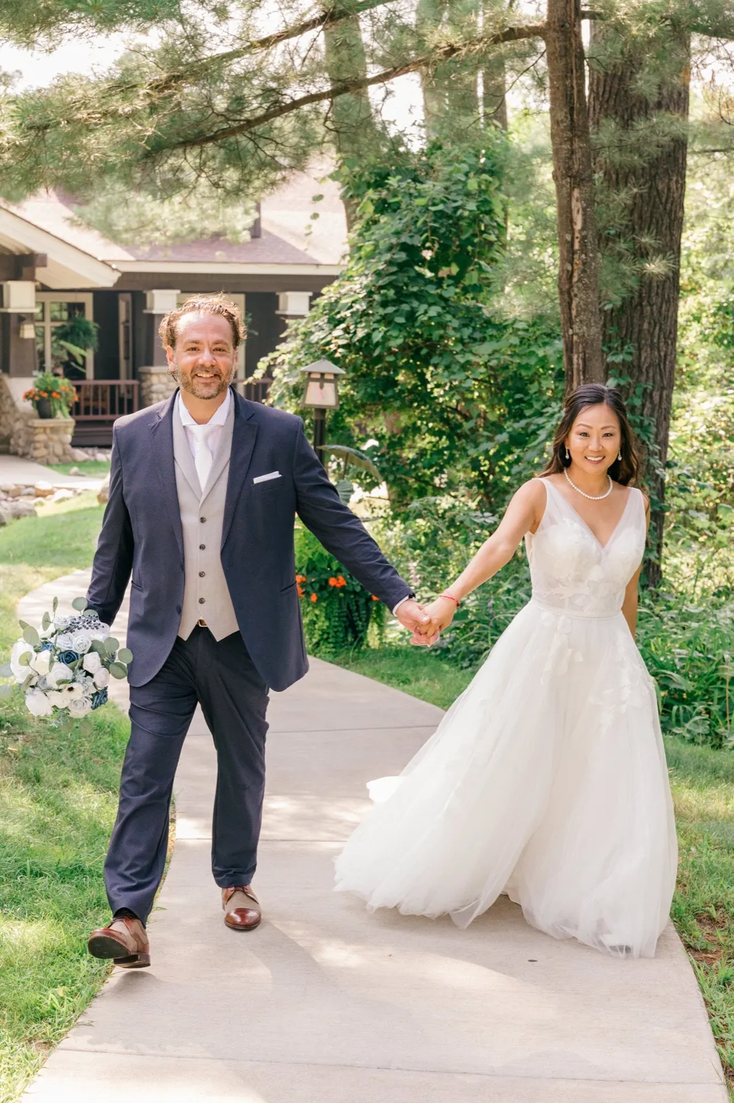 The bride and groom hold hands and walk together along a paved path in front of the lodge, surrounded by tall pines at Grand View Lodge — Tim Larsen Photography, Brainerd Lakes MN