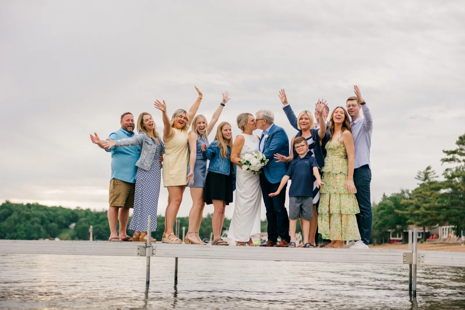 The couple shares a kiss surrounded by family with raised arms cheering on the long Grand View Lodge dock, Gull Lake stretching behind — Tim Larsen Photography, Brainerd Lakes MN