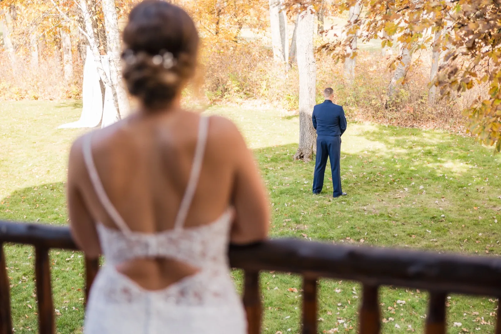 From a lodge deck, the bride stands out of focus in the foreground while the groom waits with his back turned on a sunlit autumn lawn — Tim Larsen Photography, Brainerd Lakes MN