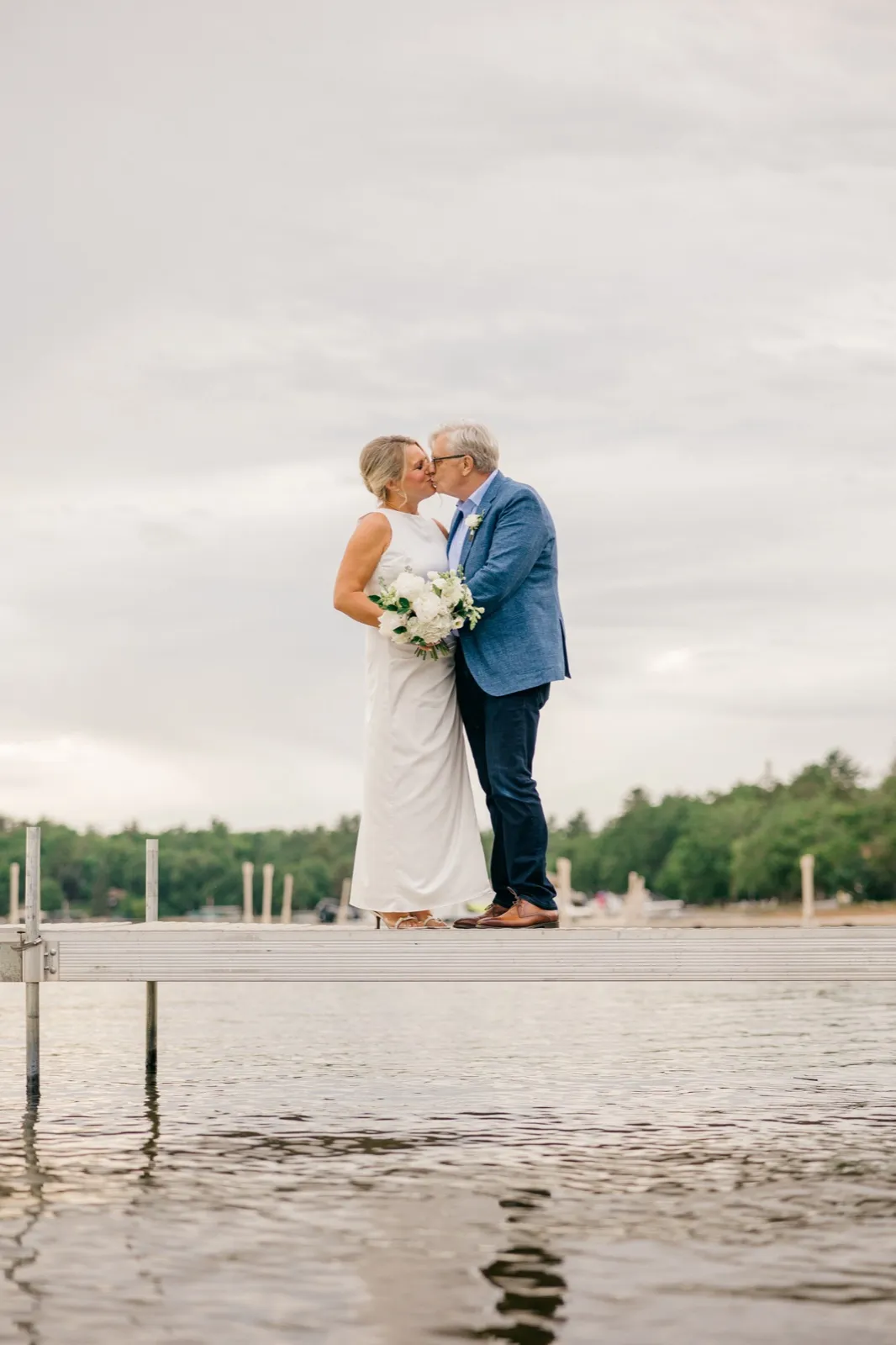 The bride and groom share a kiss at the end of the long Grand View Lodge dock over Gull Lake, the bride holding her white peony bouquet under a wide overcast sky — Tim Larsen Photography, Brainerd Lakes MN