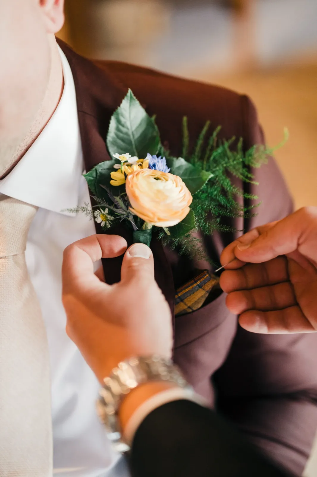 Two pairs of hands work together to pin a peach ranunculus and wildflower boutonniere onto the groom's brown suit lapel. — Tim Larsen Photography, Brainerd Lakes MN