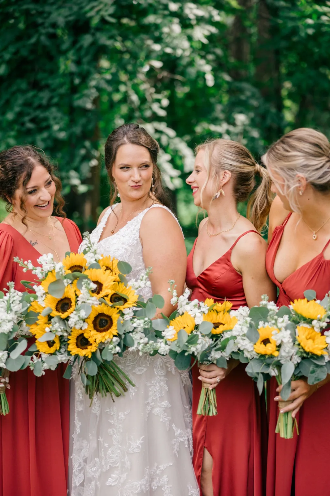 The bride makes a playful face while her bridesmaids in rust dresses laugh and hold sunflower bouquets in front of green foliage
