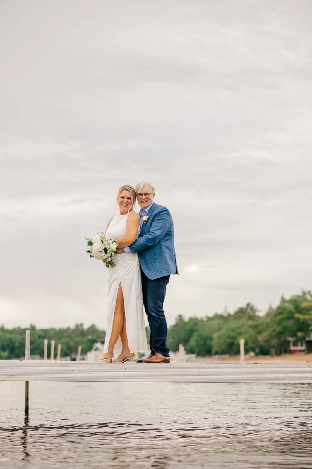 The bride and groom stand together on the wooden dock over Gull Lake, smiling toward the camera under a wide overcast June sky — Tim Larsen Photography, Brainerd Lakes MN