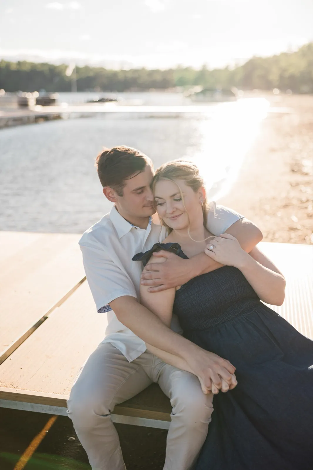 A man cradles his fiancee from behind as they sit on the Gull Lake beach at Grand View Lodge, backlit by hazy golden sun — Tim Larsen Photography