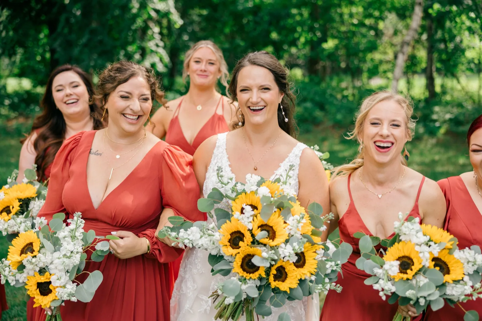 Chelsea and her bridesmaids in rust-colored dresses laugh together while holding bouquets of sunflowers and white florals on a green lawn — Tim Larsen Photography, Brainerd Lakes MN