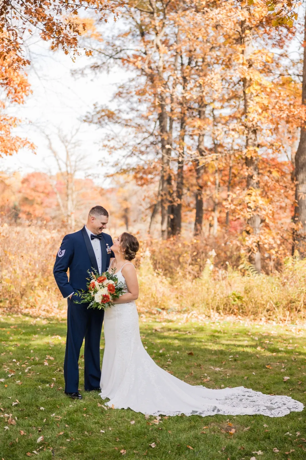 The bride in a lace-trained gown holds an autumn bouquet and looks up at the groom in his Air Force dress uniform beneath a canopy of orange fall leaves — Tim Larsen Photography, Brainerd Lakes MN