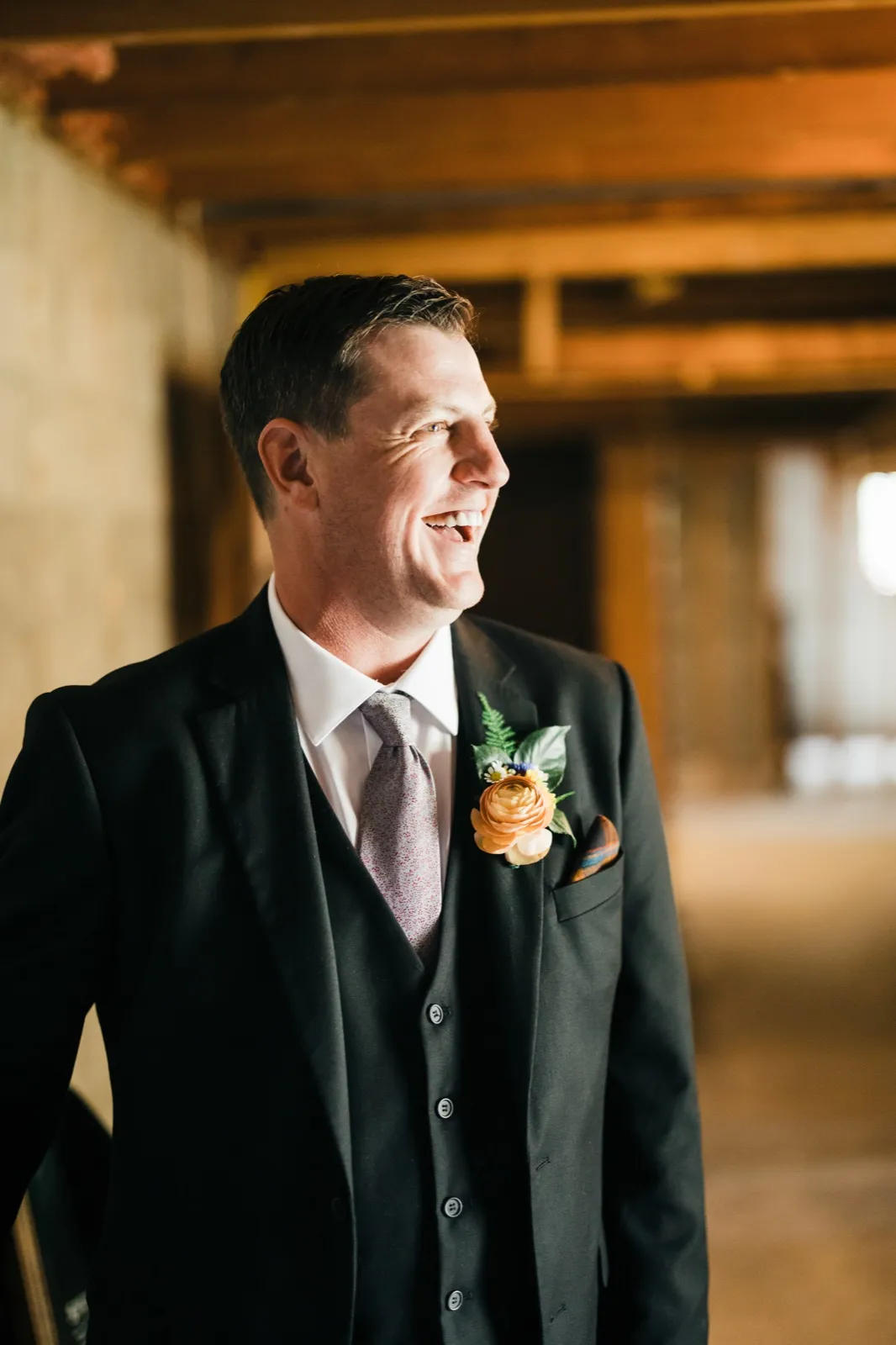 The groom in a black three-piece suit and patterned tie laughs while looking off-camera toward window light, his peach ranunculus boutonniere visible against his lapel. — Tim Larsen Photography, Brainerd Lakes MN