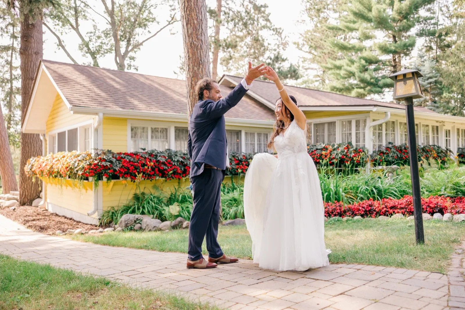 The groom twirls the bride on a paver walkway in front of a yellow cottage lined with red impatiens at Grand View Lodge — Tim Larsen Photography, Brainerd Lakes MN
