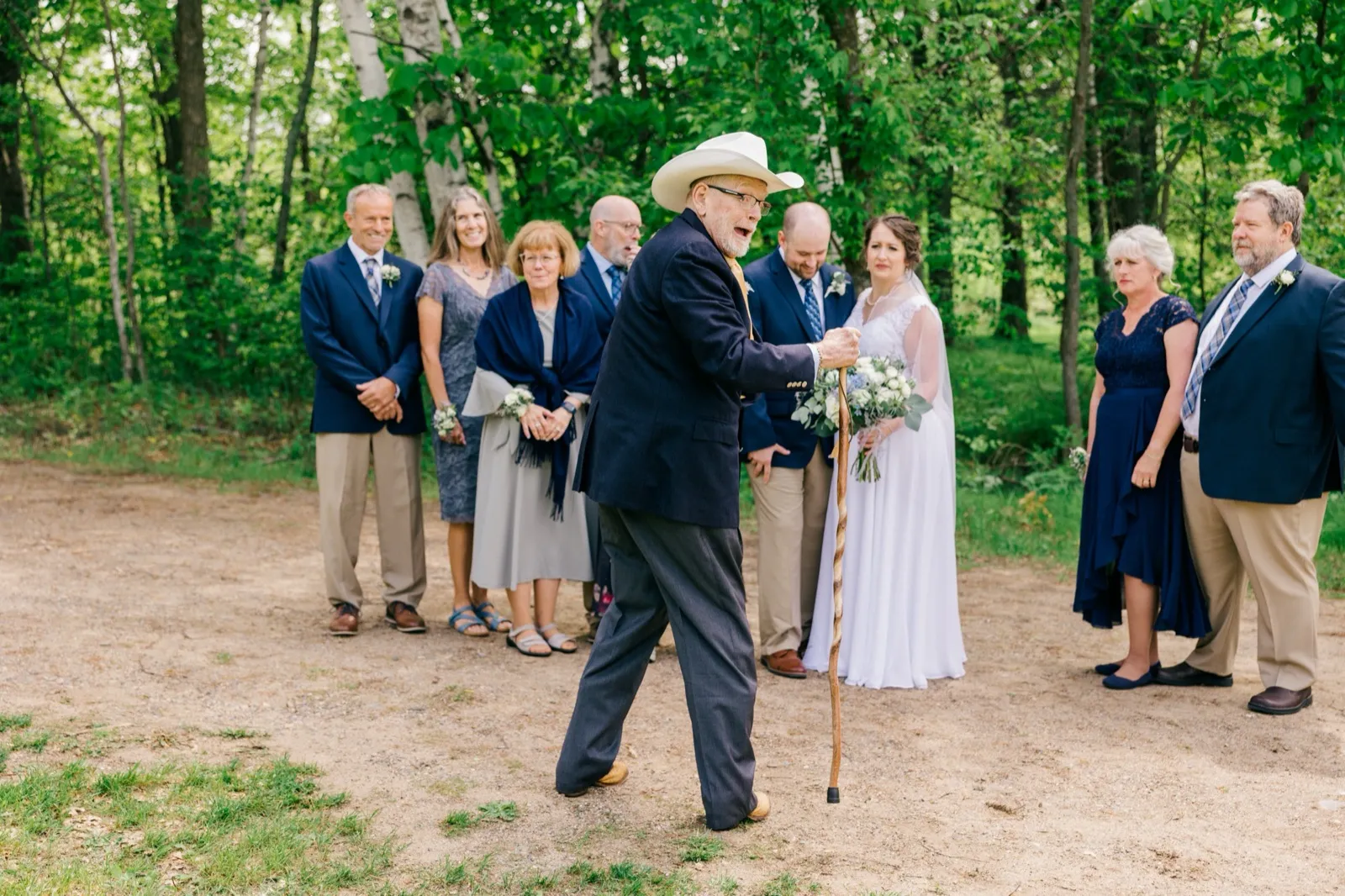 An older man in a cowboy hat walks up with a cane and a wide grin while the rest of the family waits, smiling, in a forest clearing — the kind of group a wrangler keeps moving — Tim Larsen Photography, Brainerd Lakes MN
