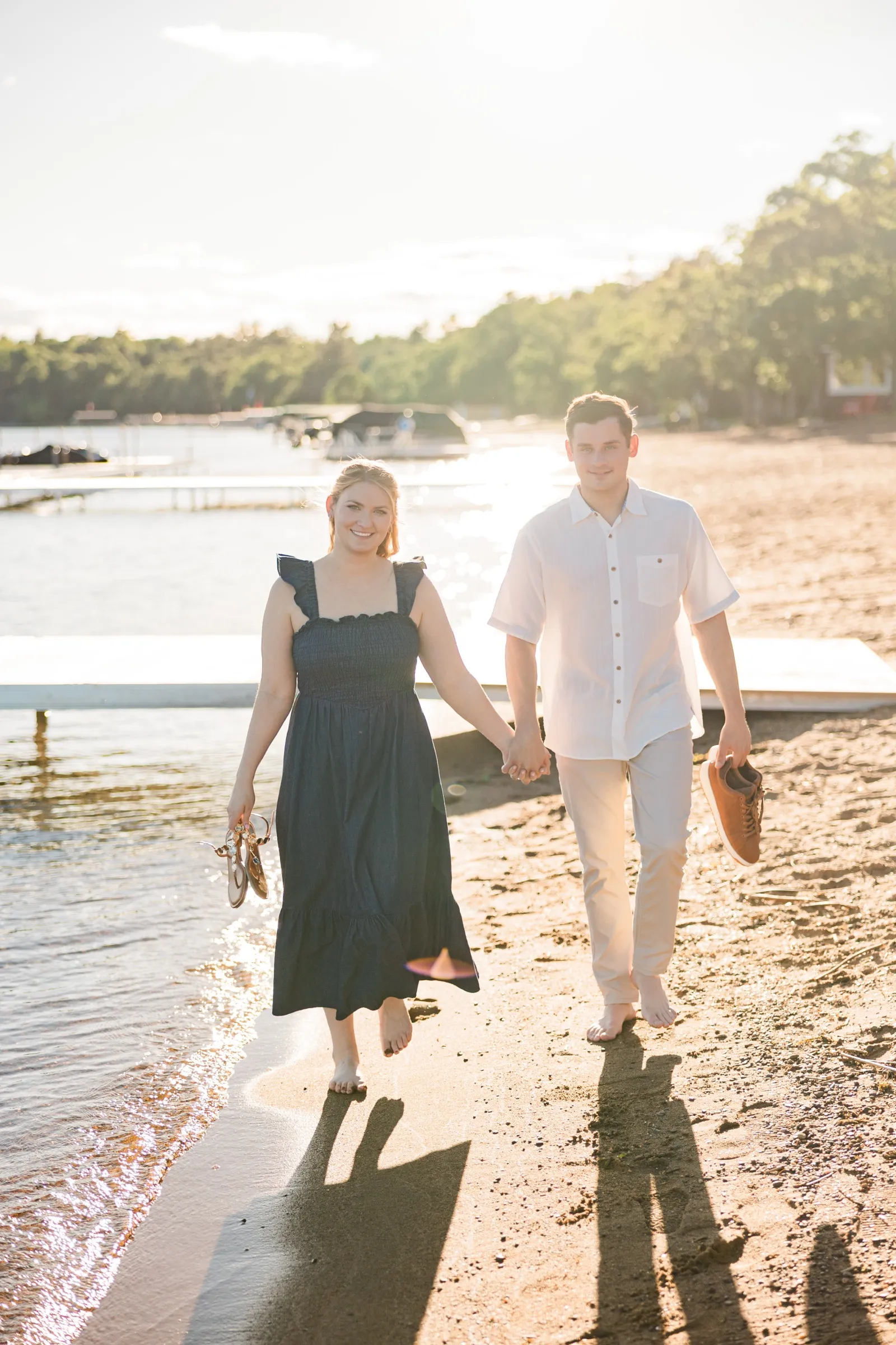 A barefoot engaged couple walk toward the camera along the Gull Lake beach, shoes in hand, backlit by the sun — Tim Larsen Photography