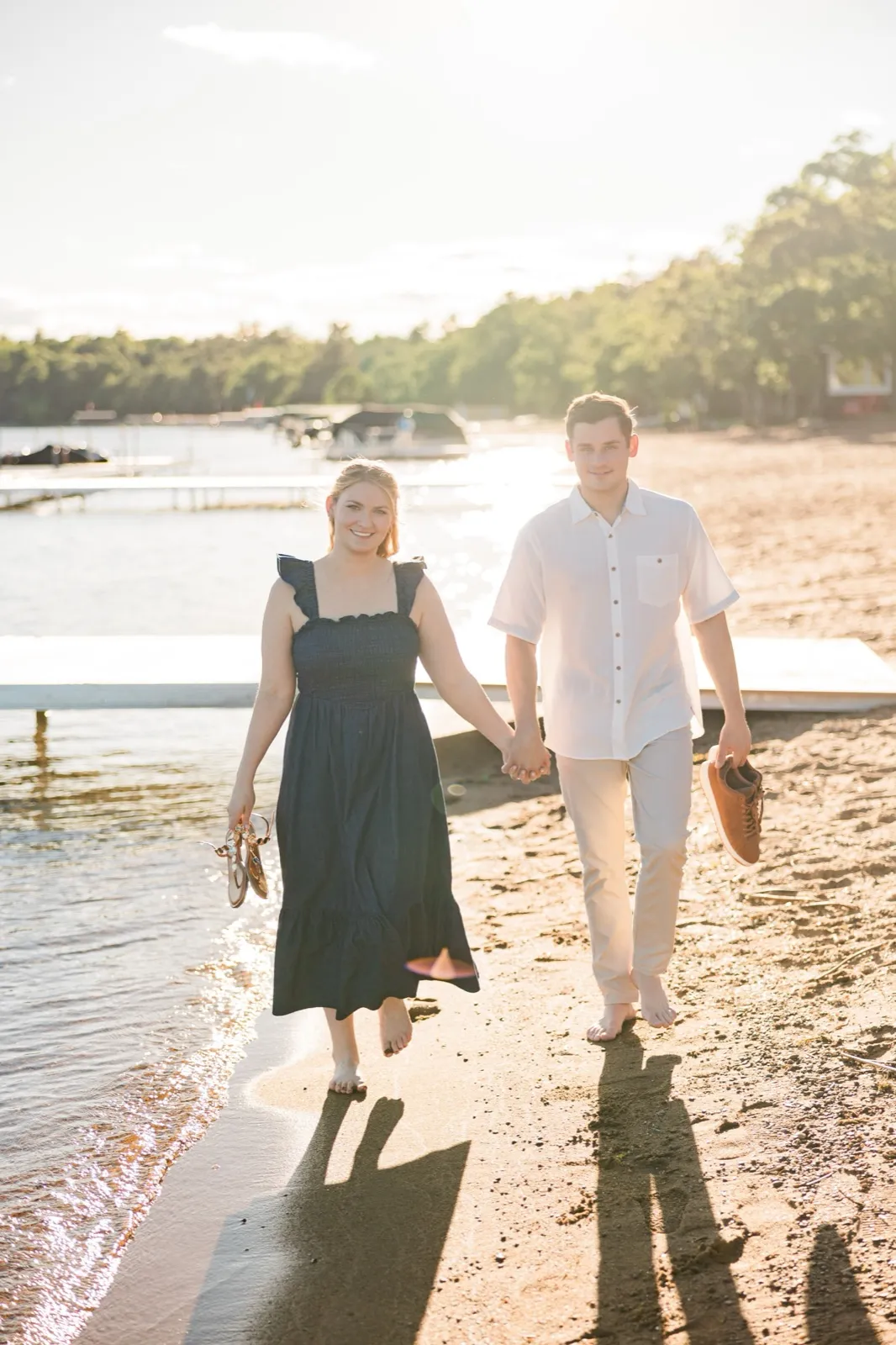 A barefoot engaged couple walk toward the camera along the Gull Lake beach, shoes in hand, backlit by the sun — Tim Larsen Photography