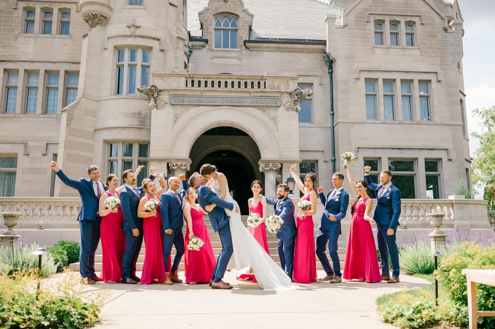 The groom dips the bride for a kiss as their bridal party in magenta dresses and navy suits cheers around them in front of the stone facade of the American Swedish Institute. — Tim Larsen Photography, Minneapolis MN
