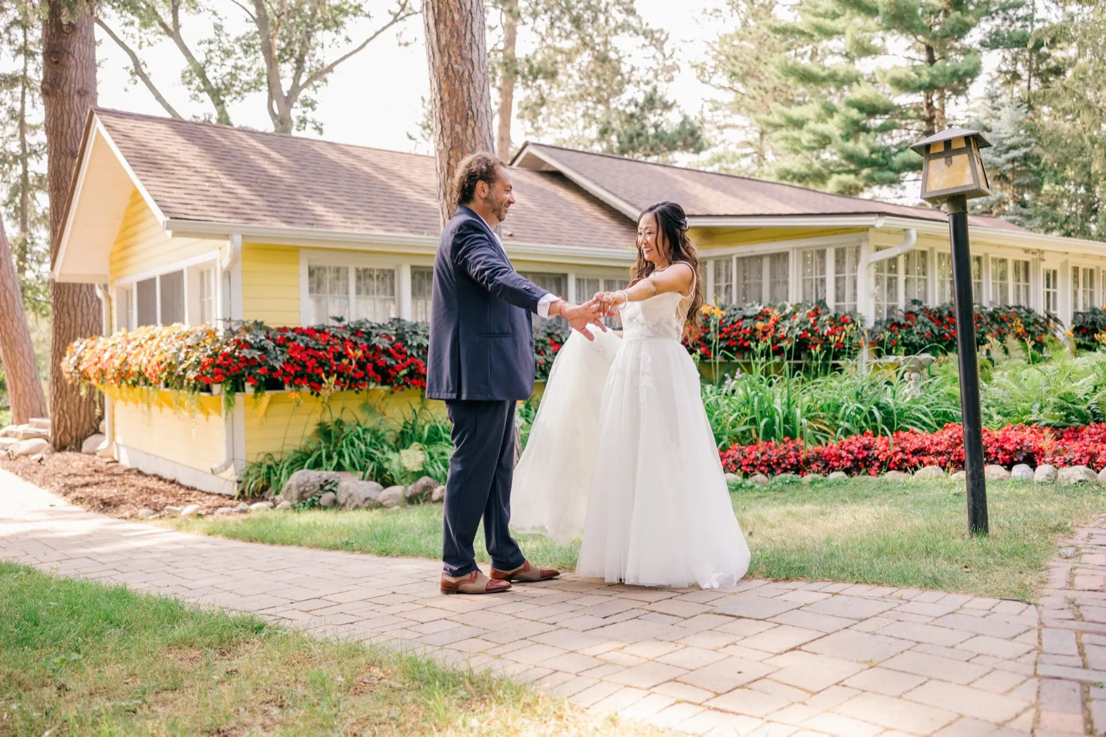 The bride and groom reach toward each other laughing on a paver path outside a yellow cottage lined with red impatiens at Grand View Lodge — Tim Larsen Photography, Brainerd Lakes MN