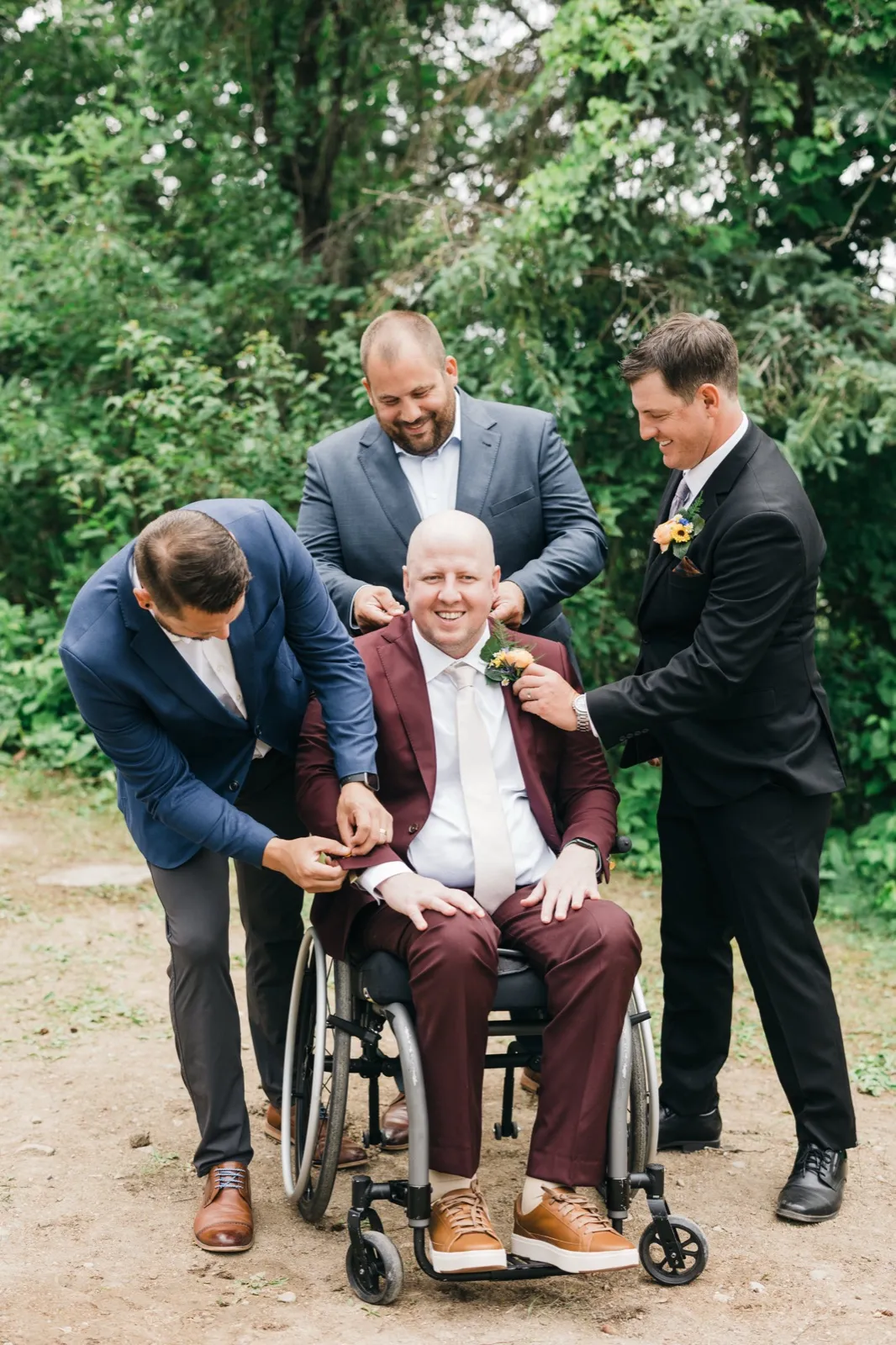 Three groomsmen in suits gather around the groom seated in a wheelchair, adjusting his boutonniere and cuff while everyone smiles on a wooded path. — Tim Larsen Photography, Brainerd Lakes MN