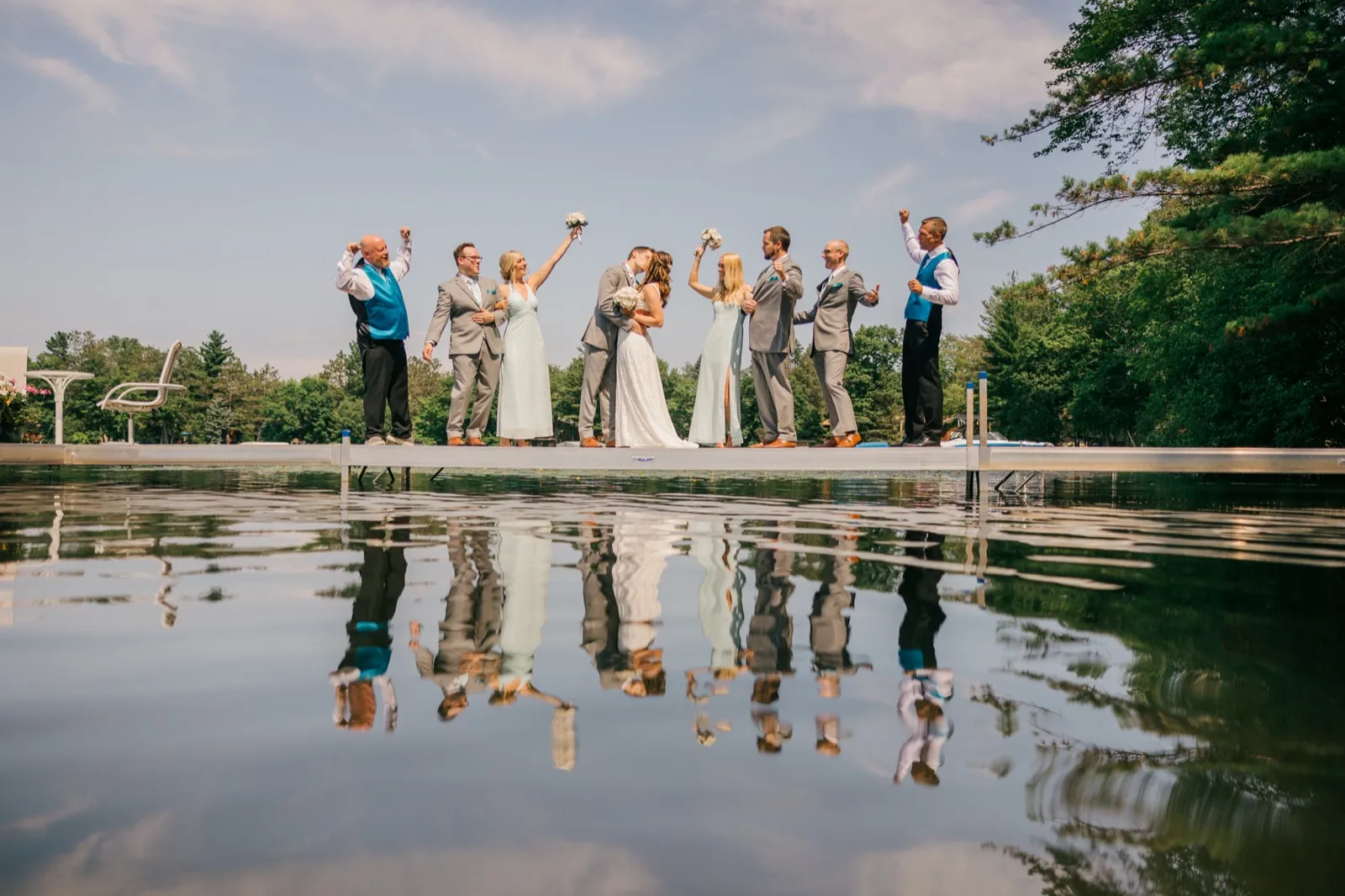 The couple kisses on a lake dock as their bridal party cheers with raised arms, all of them mirrored in glassy water below. — Tim Larsen Photography, Brainerd Lakes MN