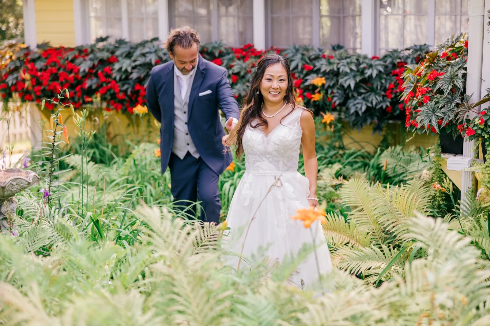 The bride smiles brightly while leading the groom by the hand through a lush garden of ferns and red blooms beside a yellow cottage at Grand View Lodge — Tim Larsen Photography, Brainerd Lakes MN