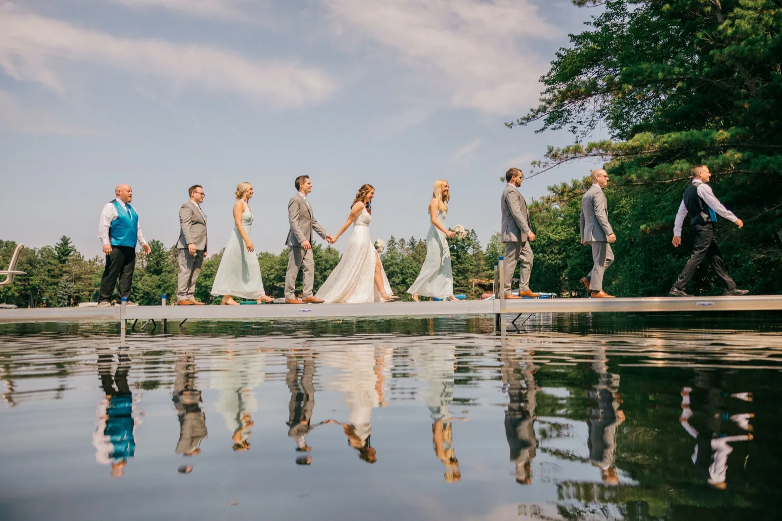 The bridal party walks single-file along a dock over a still lake, their figures mirrored in the water below. — Tim Larsen Photography, Brainerd Lakes MN