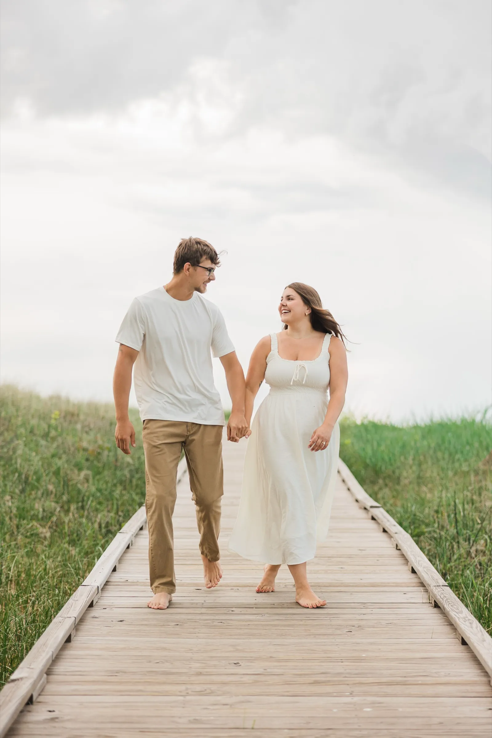 Reeden and Jacklyn walk barefoot toward the camera down the dune boardwalk, hand in hand, Jacklyn laughing — Tim Larsen Photography