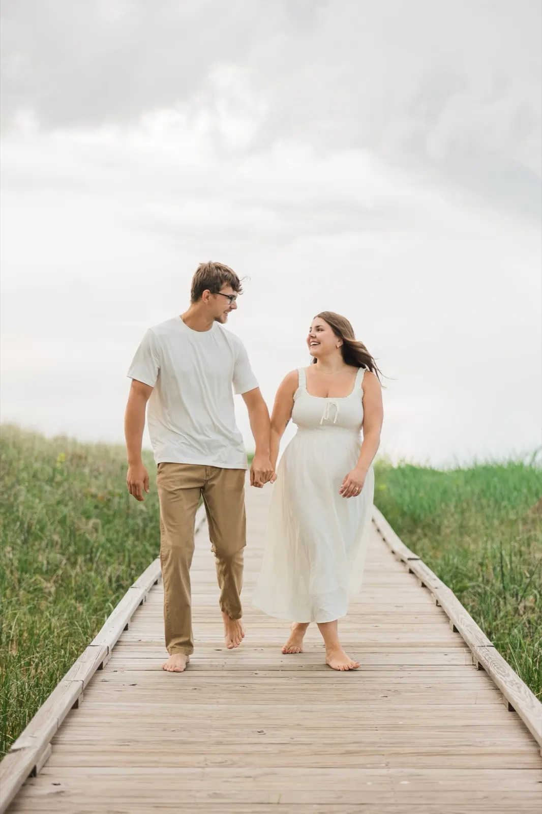 Reeden and Jacklyn walk barefoot toward the camera down the dune boardwalk, hand in hand, Jacklyn laughing — Tim Larsen Photography